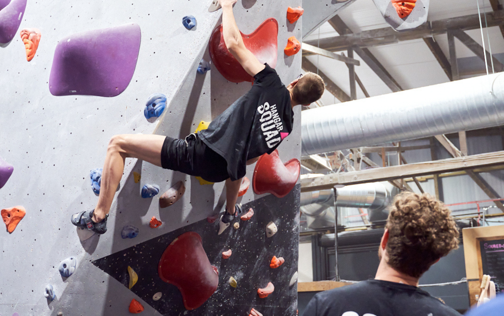 A Hangar Squad climber attempts a competition bloc under the watchful eye of Head Coach Sam Owens.